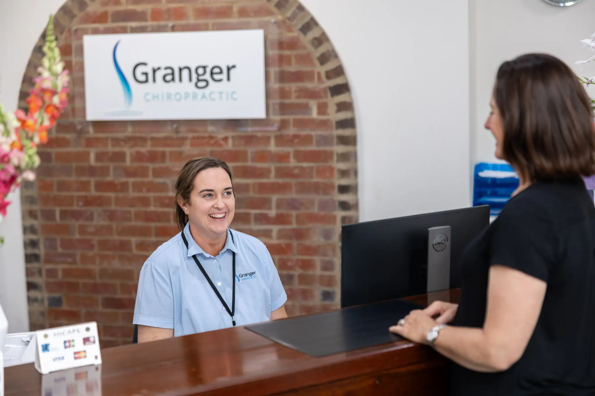 Receptionist greeting visitor at chiropractic clinic desk.